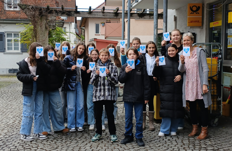 Klasse steht mit Lehrerin auf dem Markplatz und sie heben Zettel mit blauen Herzen